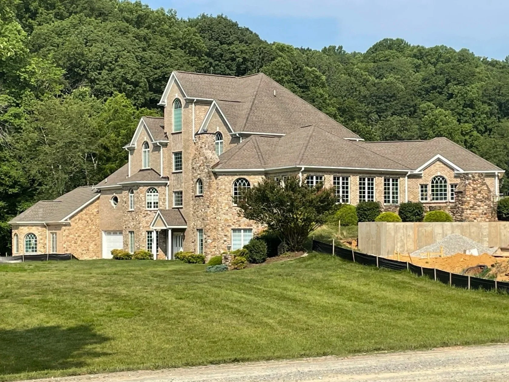A large house with a stone exterior, multiple gabled roofs, and arched windows, surrounded by landscaped green lawn and trees, with a wooded hillside in the background.