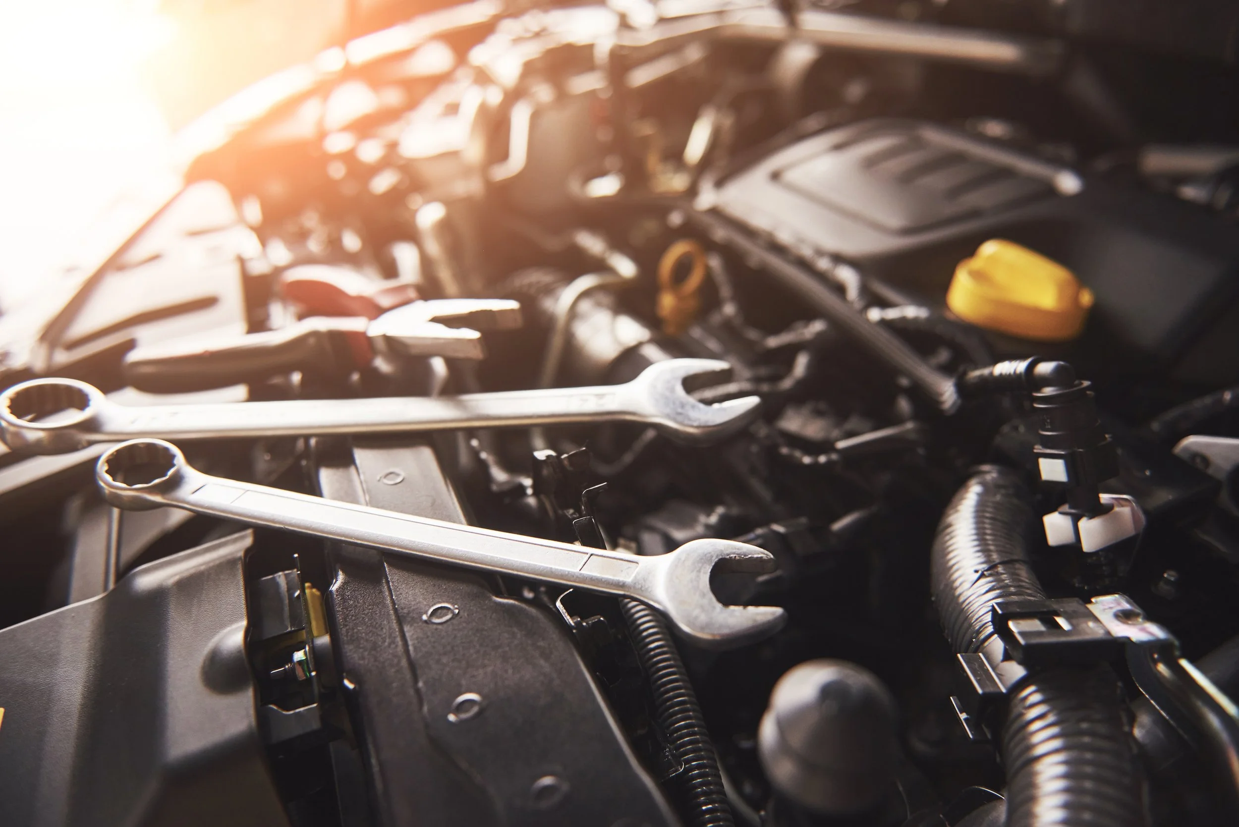 Close-up of a car engine with various mechanical parts like nuts, bolts, hoses, and a yellow cap, illuminated by sunlight.