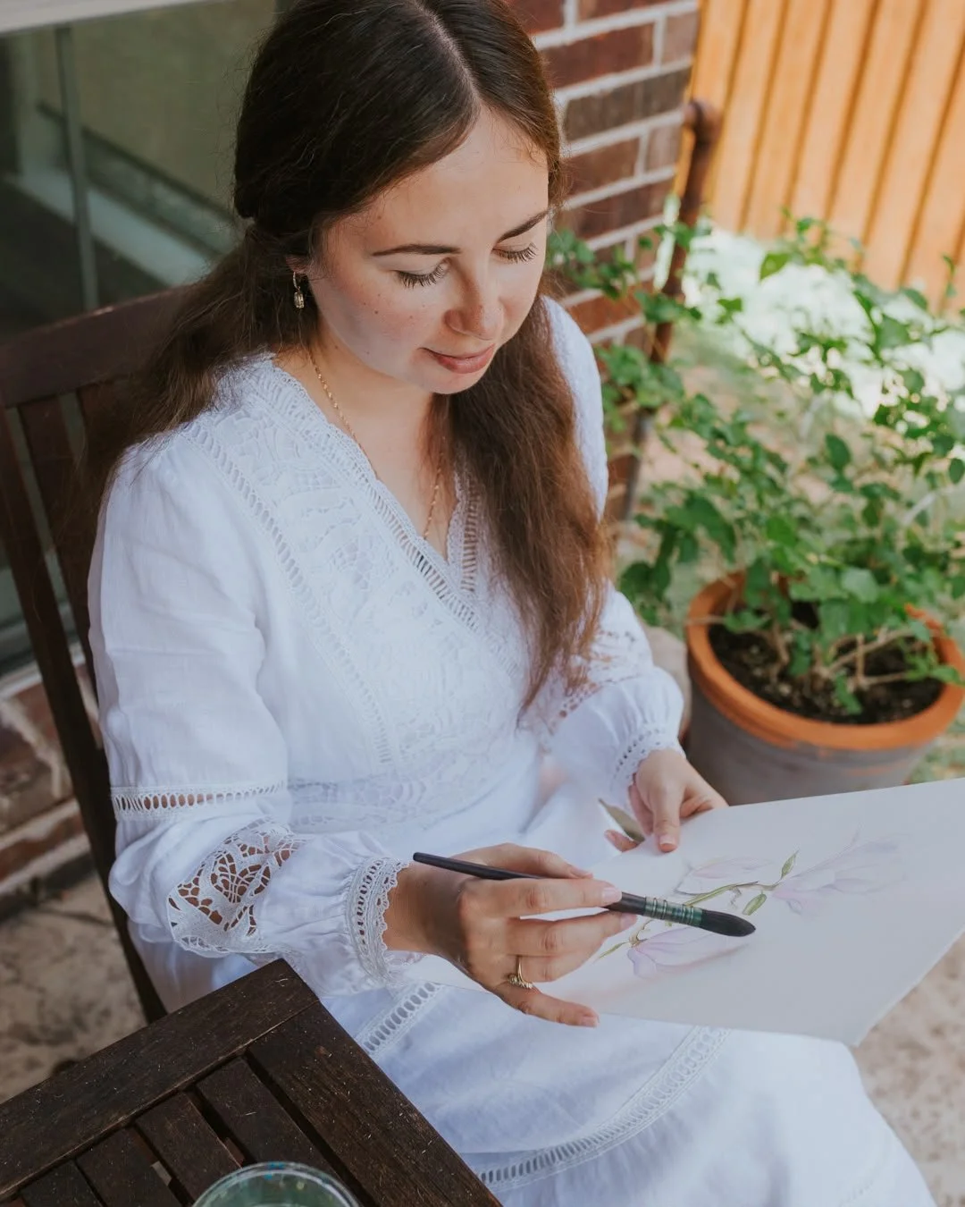 Woman in a white dress sitting outdoors, painting a floral watercolor on paper, with a potted plant and brick wall in the background.