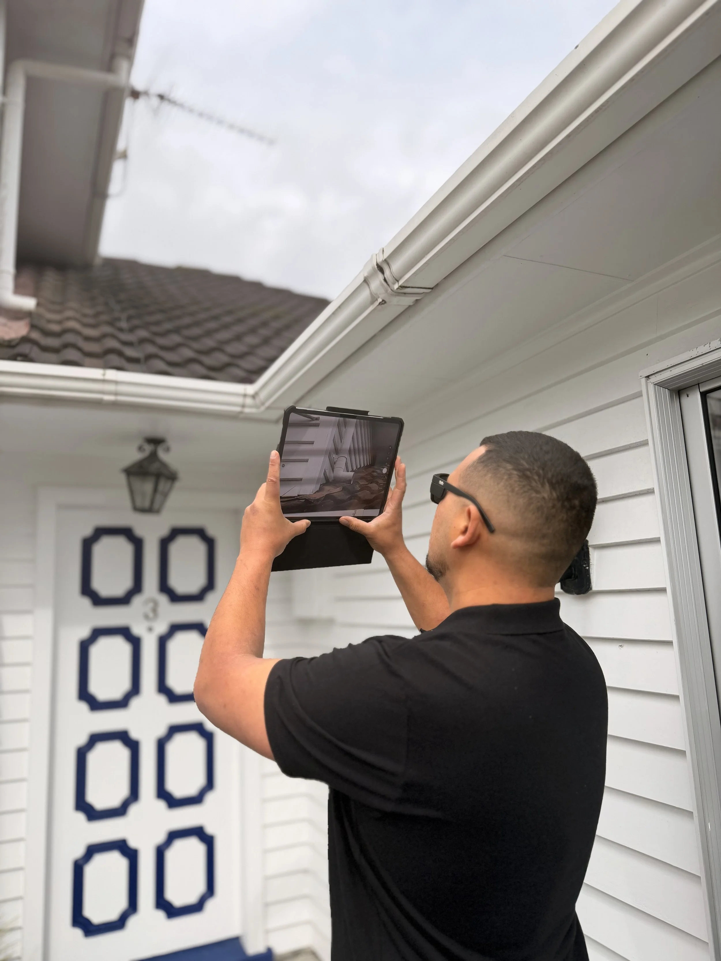 A man wearing glasses is taking a photo of a house's exterior wall and roof using a tablet. The house has white siding, a white gutter, and a door with blue geometric designs. A lantern is mounted on the wall next to the door, and a large sign with the number 00 is also visible.