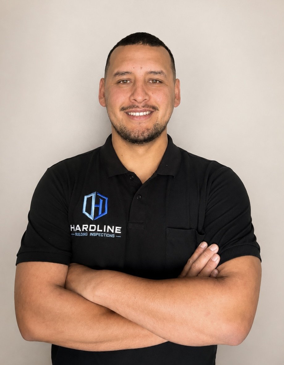 Man in black polo shirt with 'Hardline Building Inspections' logo, smiling with crossed arms, standing against a plain white wall.