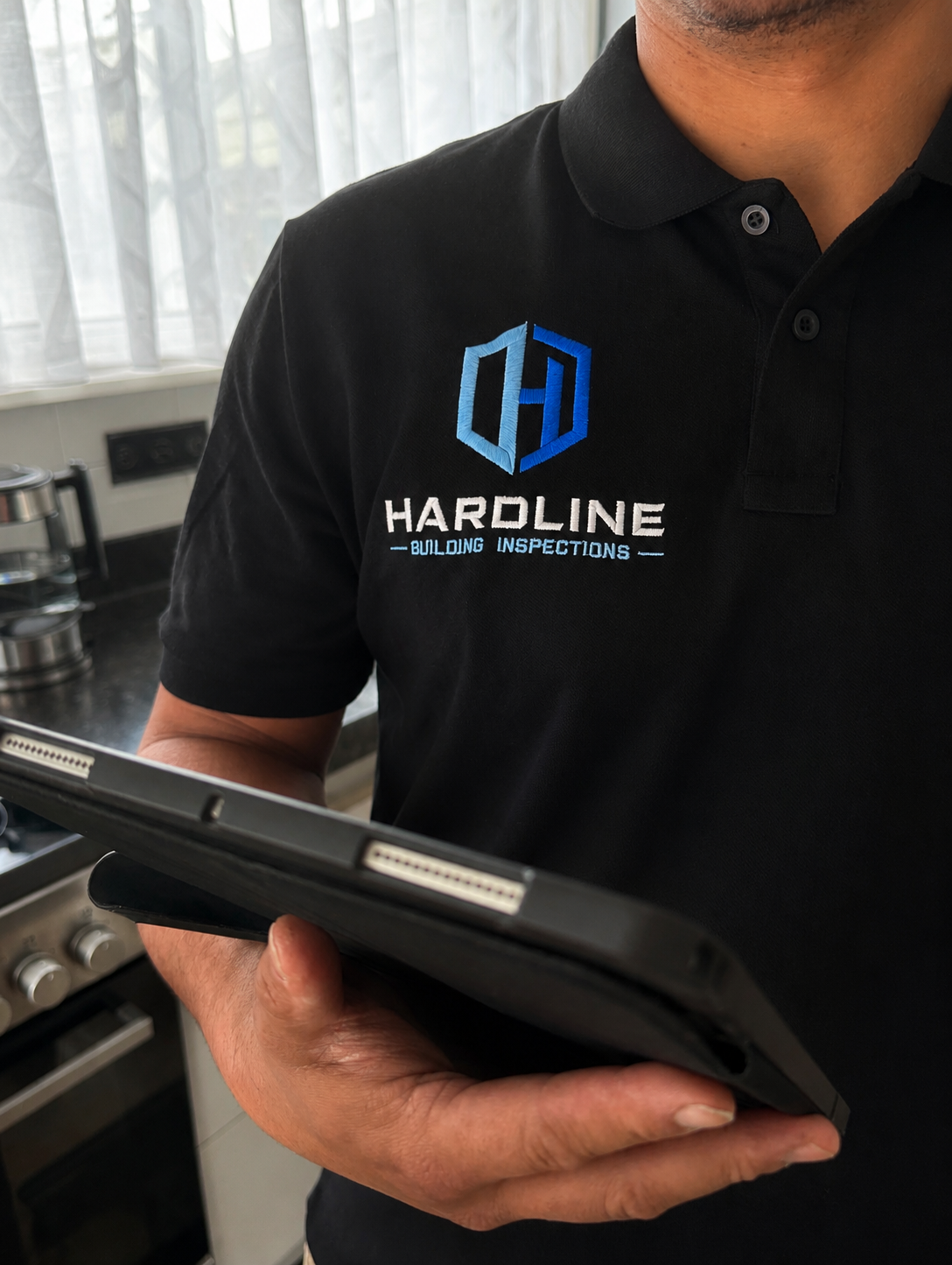A person wearing a black polo shirt with the logo and text of Hardline Building Inspections, holding a tablet device in a kitchen setting.