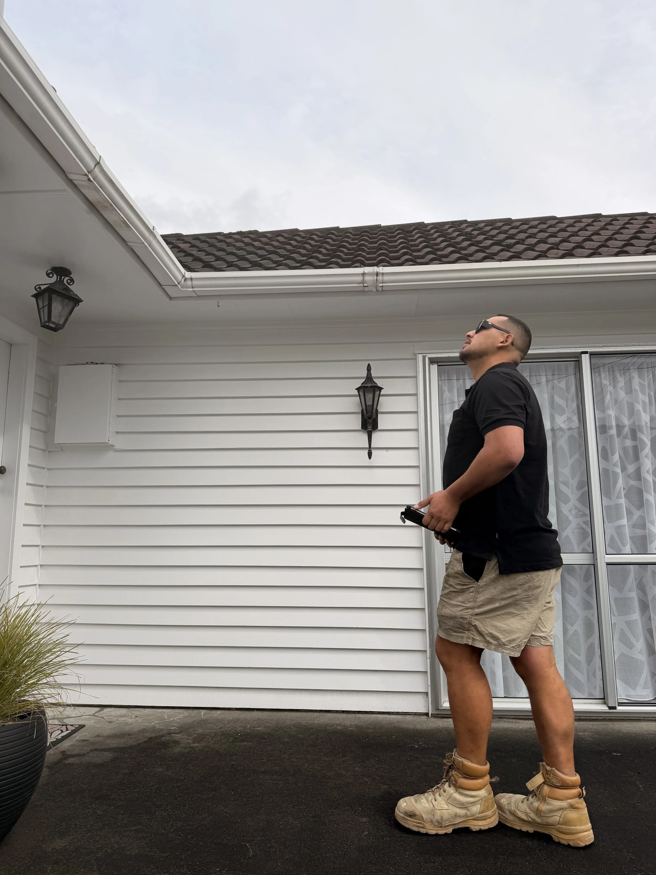 A man is standing on a concrete surface in front of a white house with black lamps, wearing oversized tan work boots, tan shorts, a black shirt, sunglasses, and holding a remote control, looking up at the sky.
