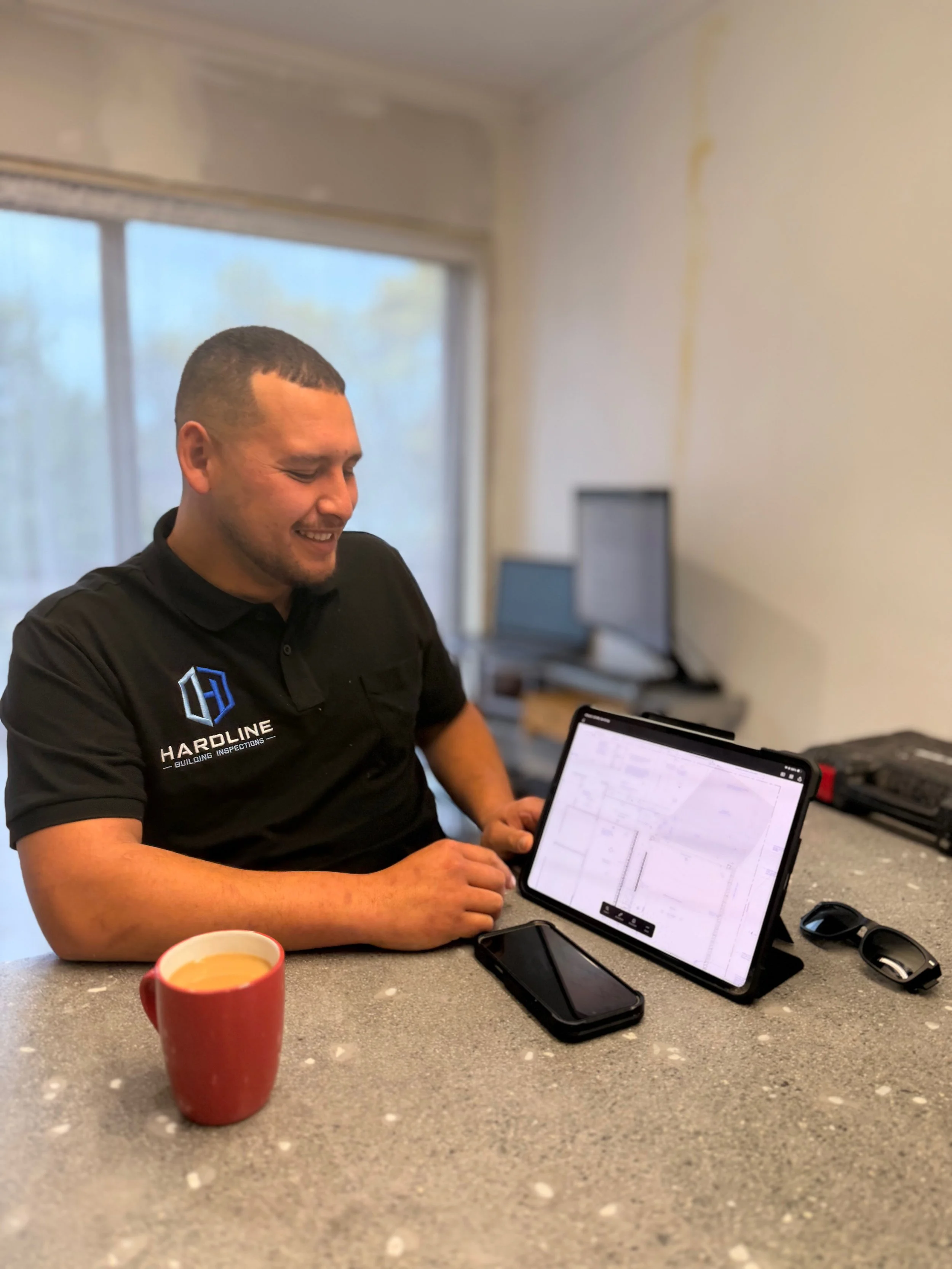 A smiling man in a black shirt sitting at a gray speckled countertop with a red coffee mug, a tablet displaying architectural plans, a smartphone, and sunglasses.