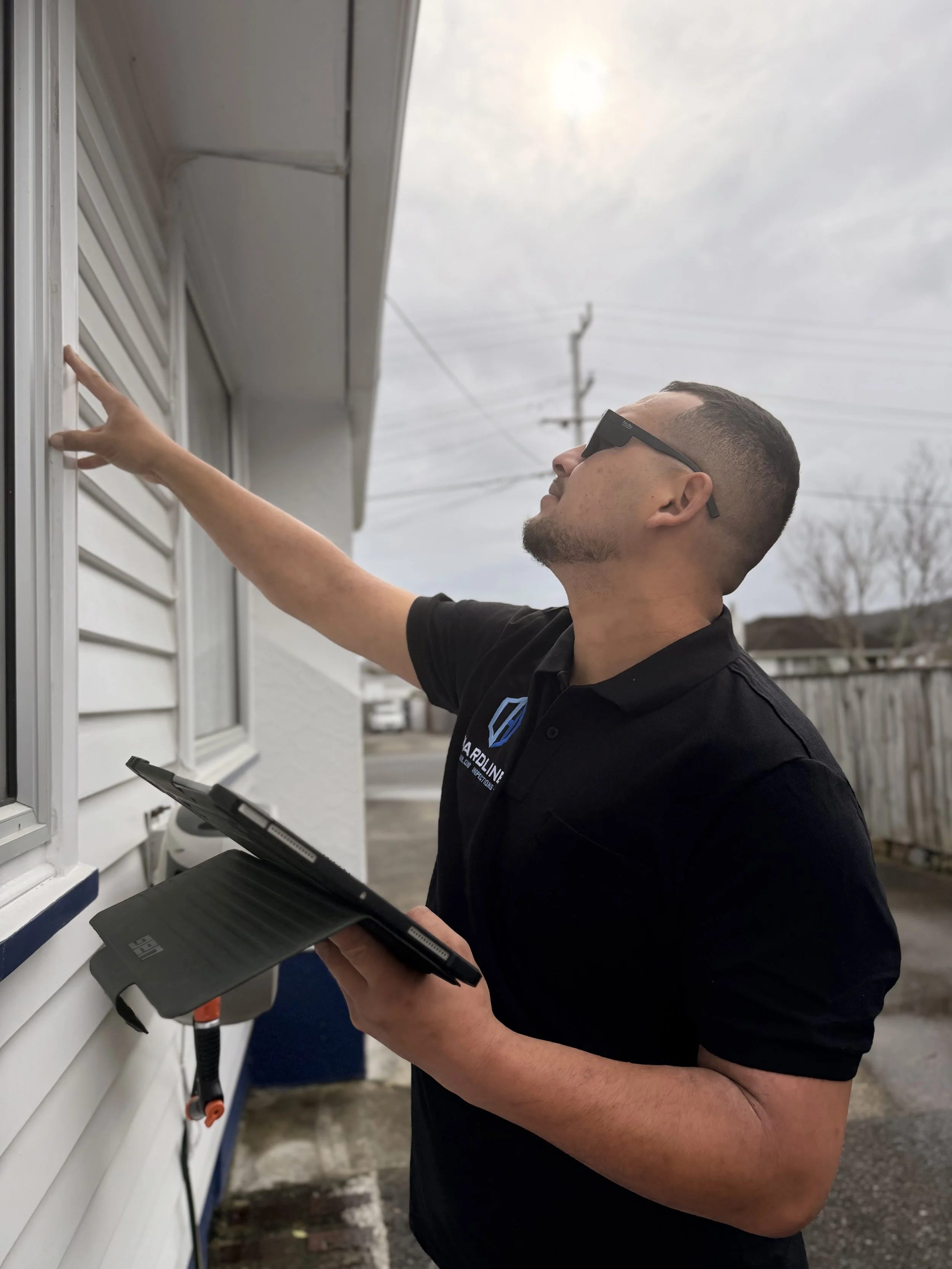 A man wearing sunglasses and a black shirt is installing or repairing a satellite dish on the side of a house, holding a tablet in one hand and reaching with the other.