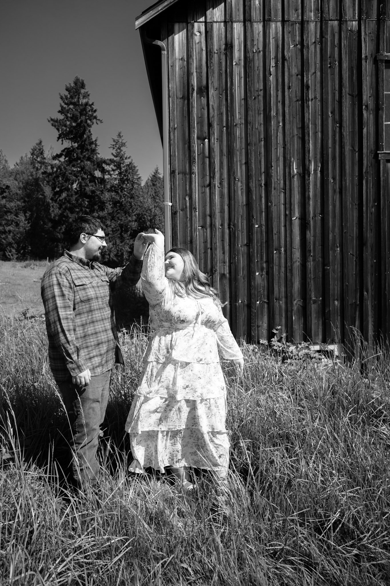 A man and a woman dance together outdoors in front of a wooden building, with trees in the background, on a sunny day.