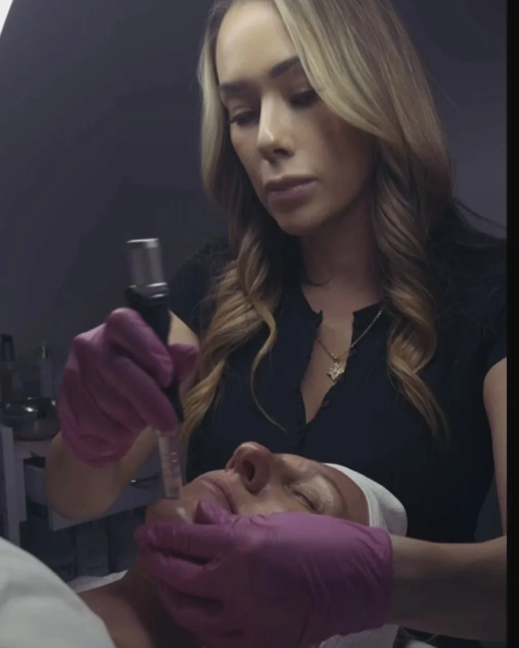 A woman with long blonde hair and a black top is performing a cosmetic procedure on a woman lying down with her eyes closed. The woman performing the procedure wears purple gloves and is using a syringe.