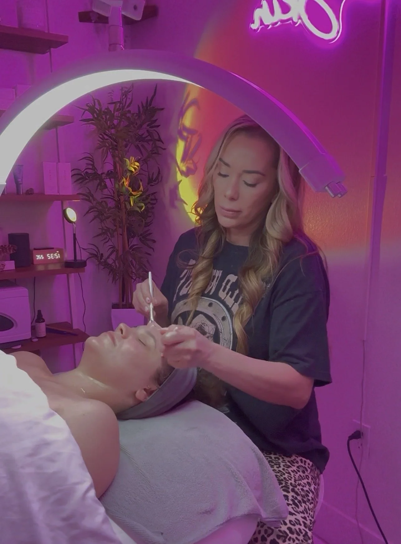 A woman receives a facial treatment with a beautician in a salon decorated with pink neon lights, a neon sign, and a potted plant, with the woman lying down and the beautician applying a facial mask or treatment.