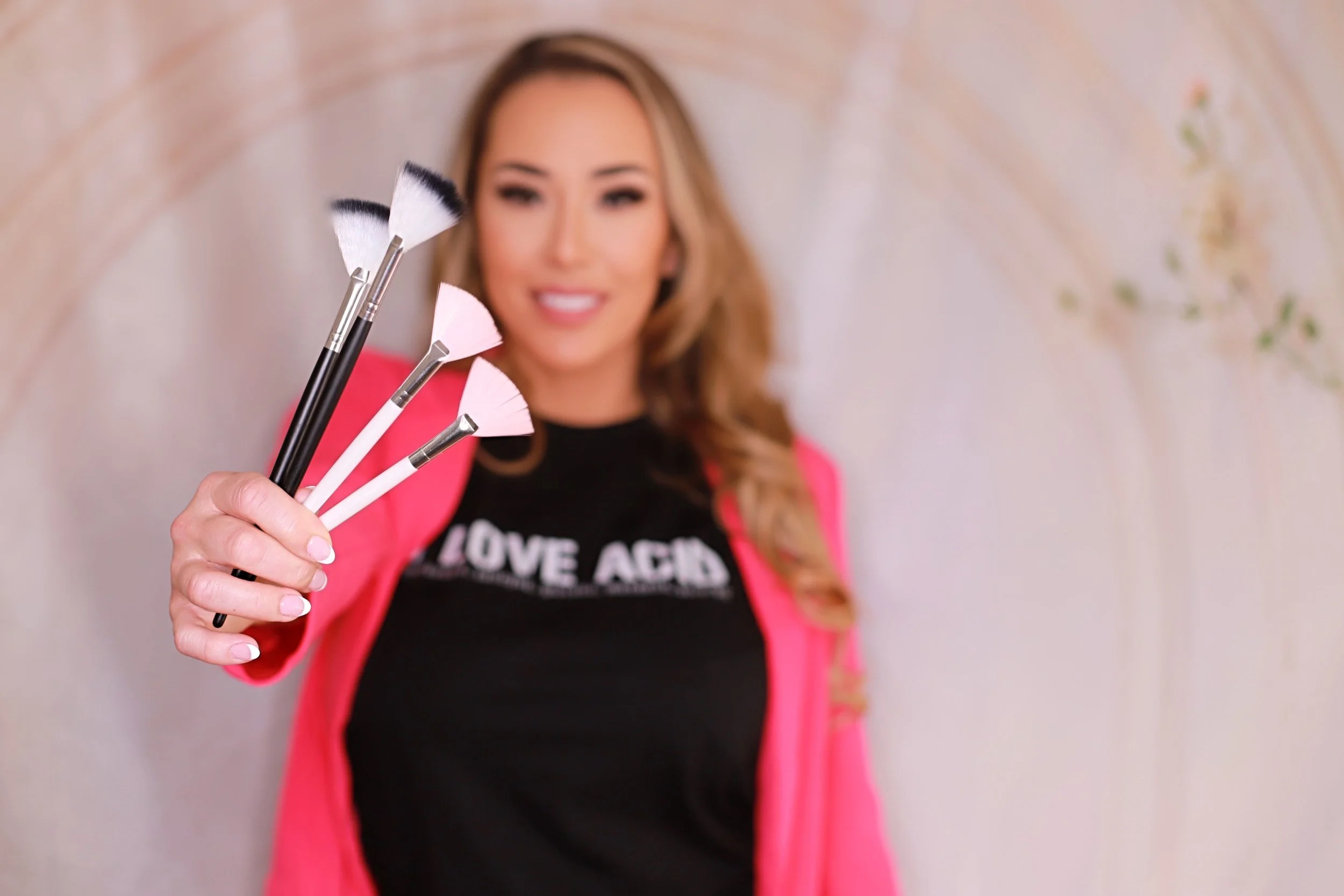 A woman is holding makeup brushes in front of her, with blurred background and a smile on her face.