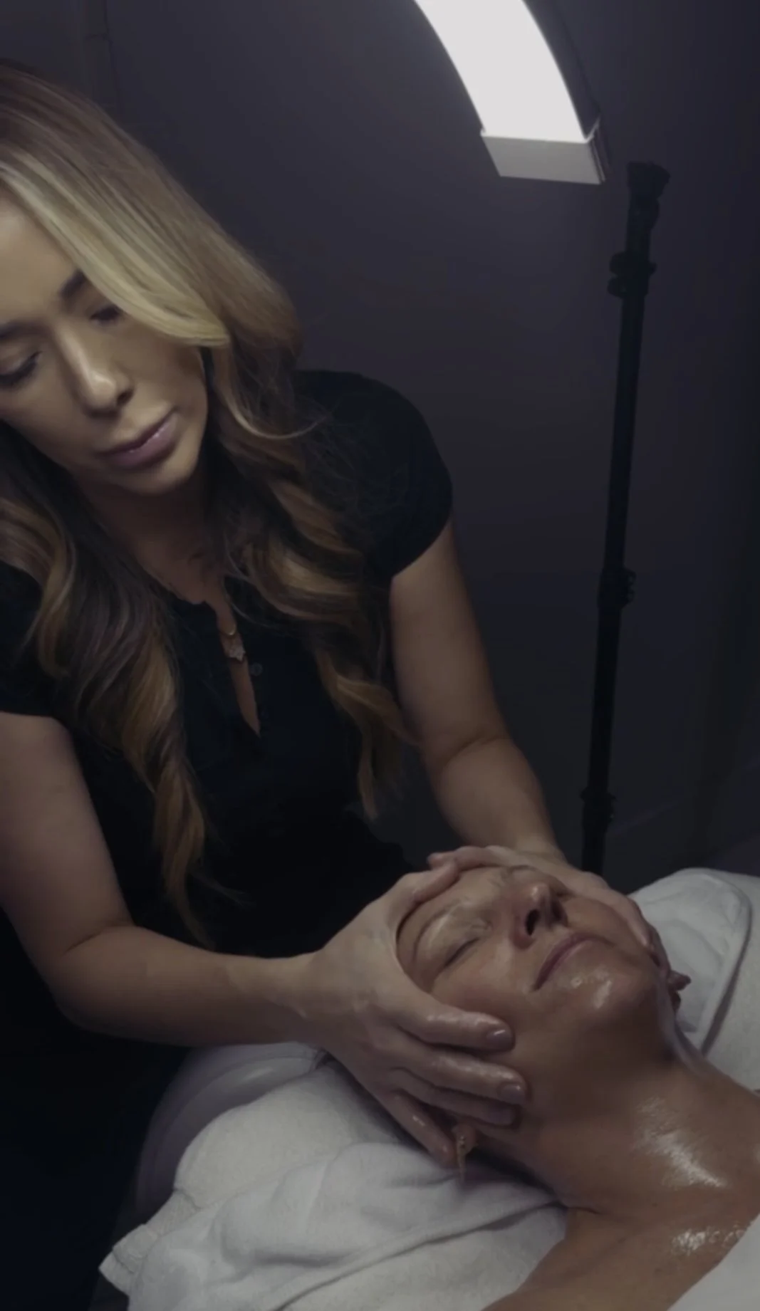 A woman receiving a facial massage from a skincare professional in a clinical setting, with a professional lighting setup overhead.