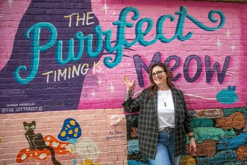 Beth McHale standing in front of a hand-painted mural for The Purrfect Meow