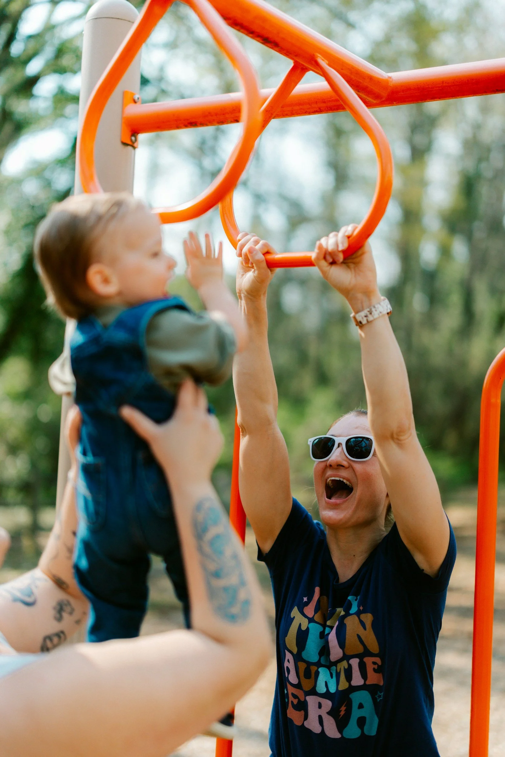 A woman wearing white sunglasses and a dark T-shirt is helping a young boy hang from an orange jungle gym bar outdoors during daytime. The boy, who has short hair and tattoos on his arm, is gripping the bar with both hands. The background features blurred trees and natural scenery.