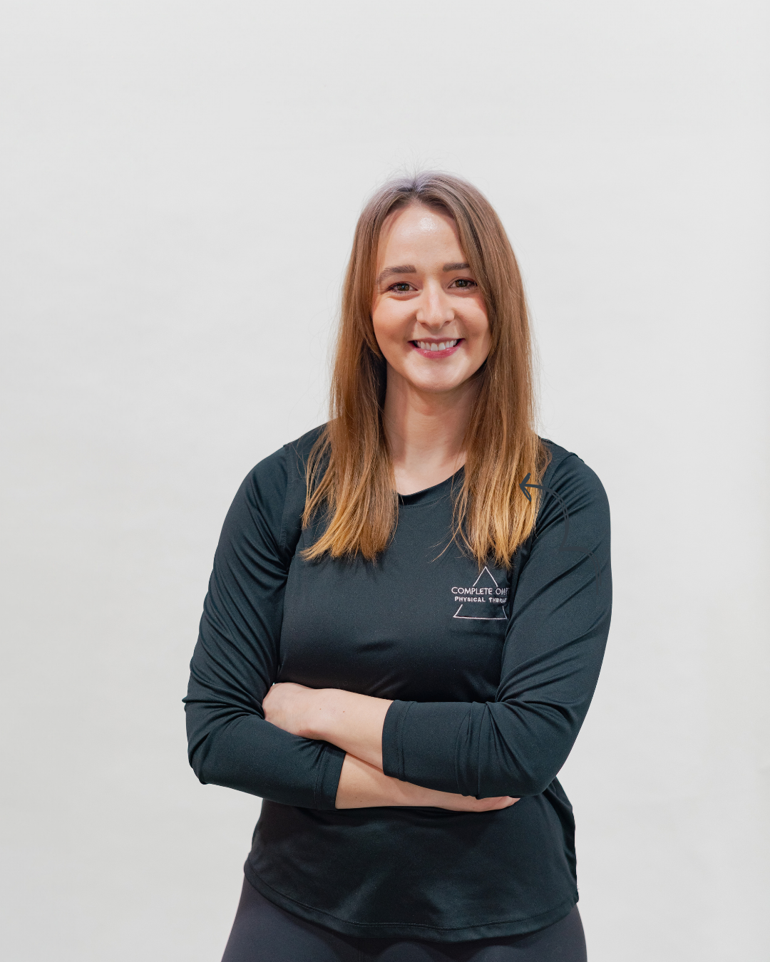 A smiling woman with long, light brown hair, wearing a black athletic shirt with the logo of 'Complete One Physical Therapy' on the chest, standing with arms crossed against a plain white background.