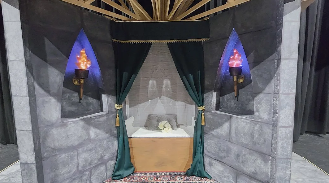 A four-poster bed with dark green curtains, flanked by stone wall alcoves with torch-like sconces emitting purple flames.