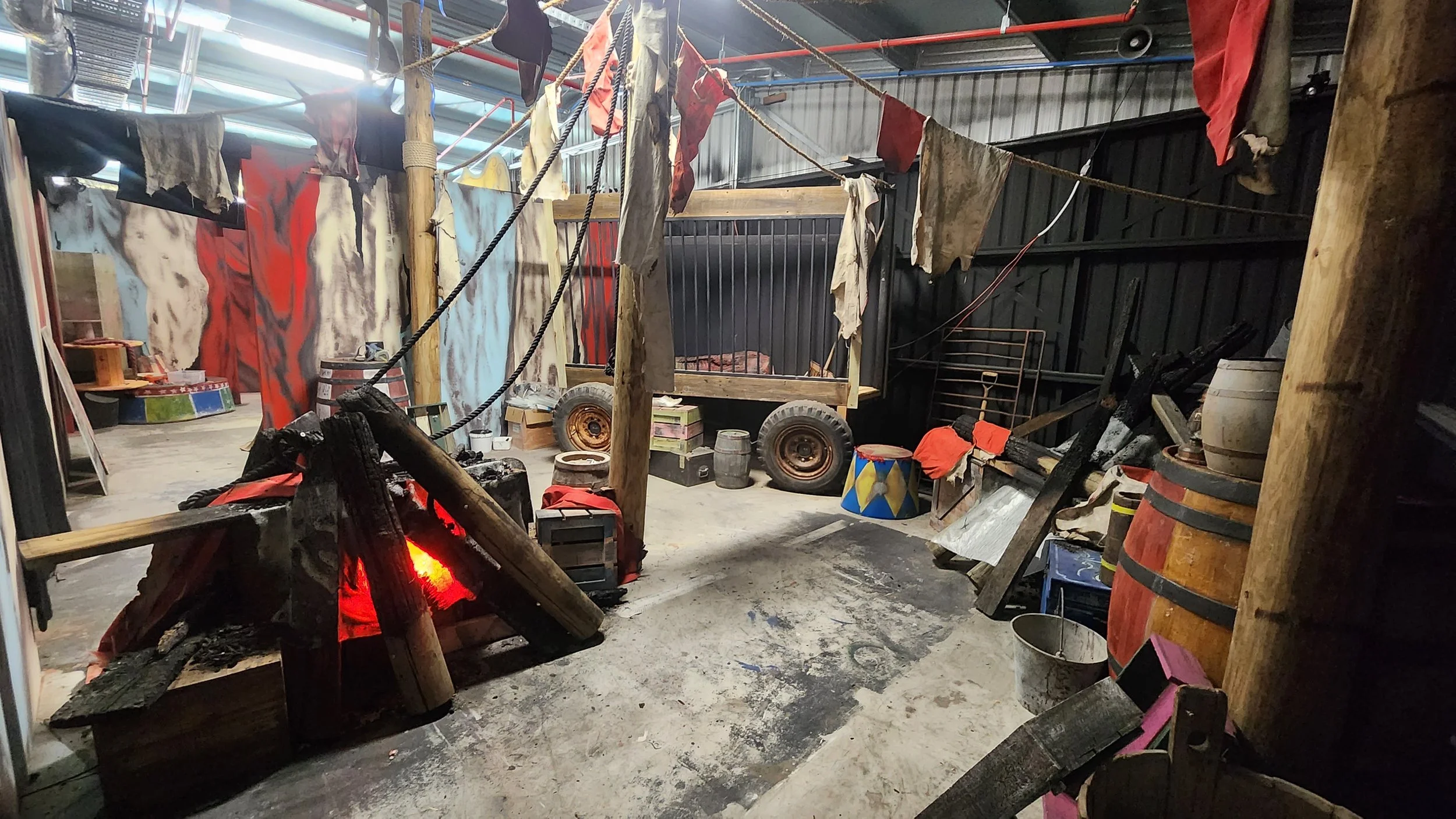 Inside a workshop or warehouse with wooden beams and ropes hanging from the ceiling, displaying various painted and weathered cloth banners or flags. There are barrels, a wooden cart, and various miscellaneous items scattered around, giving an industrial or rustic vibe.