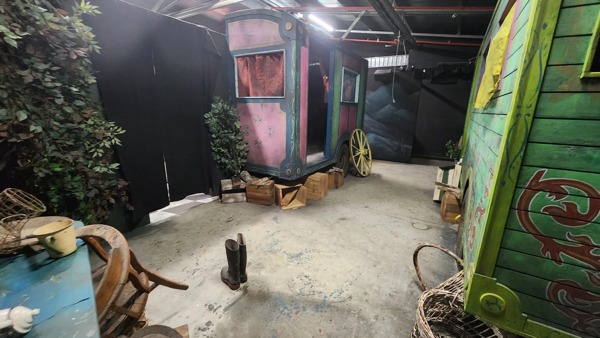 An indoor space with a vintage-style pink and blue painted cart, plants, and wooden crates, surrounded by various objects including a chair, a green painted wooden surface, and a pair of rubber boots on the floor.