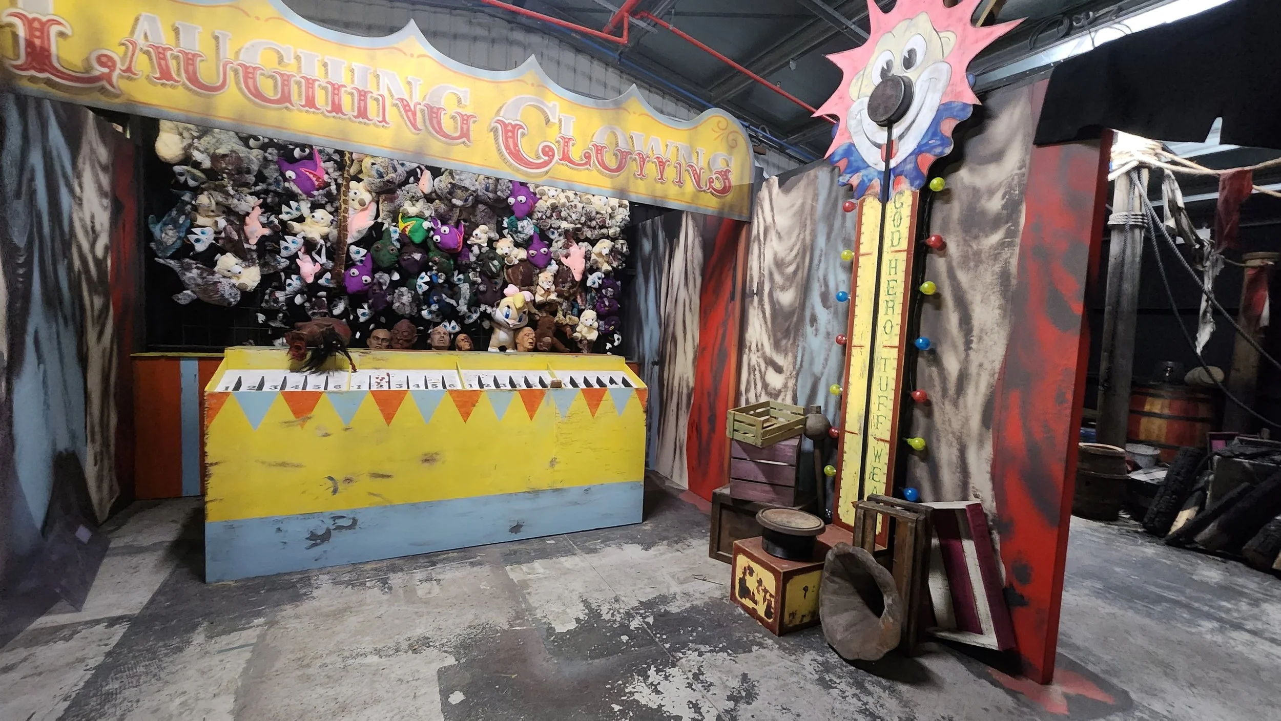 Carnival game booth featuring plush panda and unicorn stuffed animals on a wall, a yellow wooden box with numbered slots for game play, and a wooden top hat on the floor. Colorful signs and a clown face decoration with a thermometer-like scale indicating a 'You Win' zone are also visible.