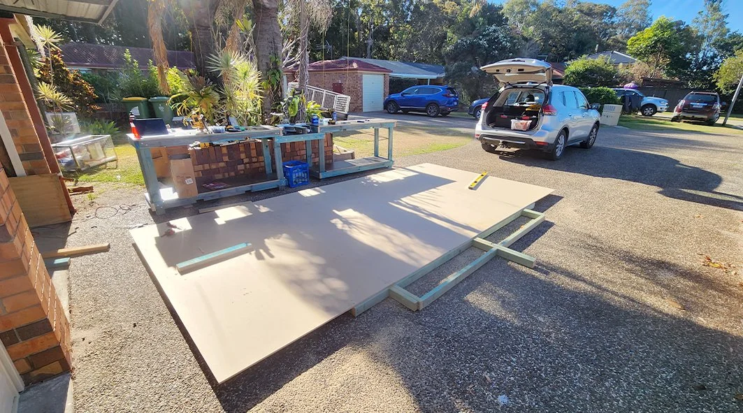 Garage or driveway area under construction with large installed white panels, tools, and a table with gardening supplies. A vehicle with an open trunk is parked nearby, and other cars are visible in the background.