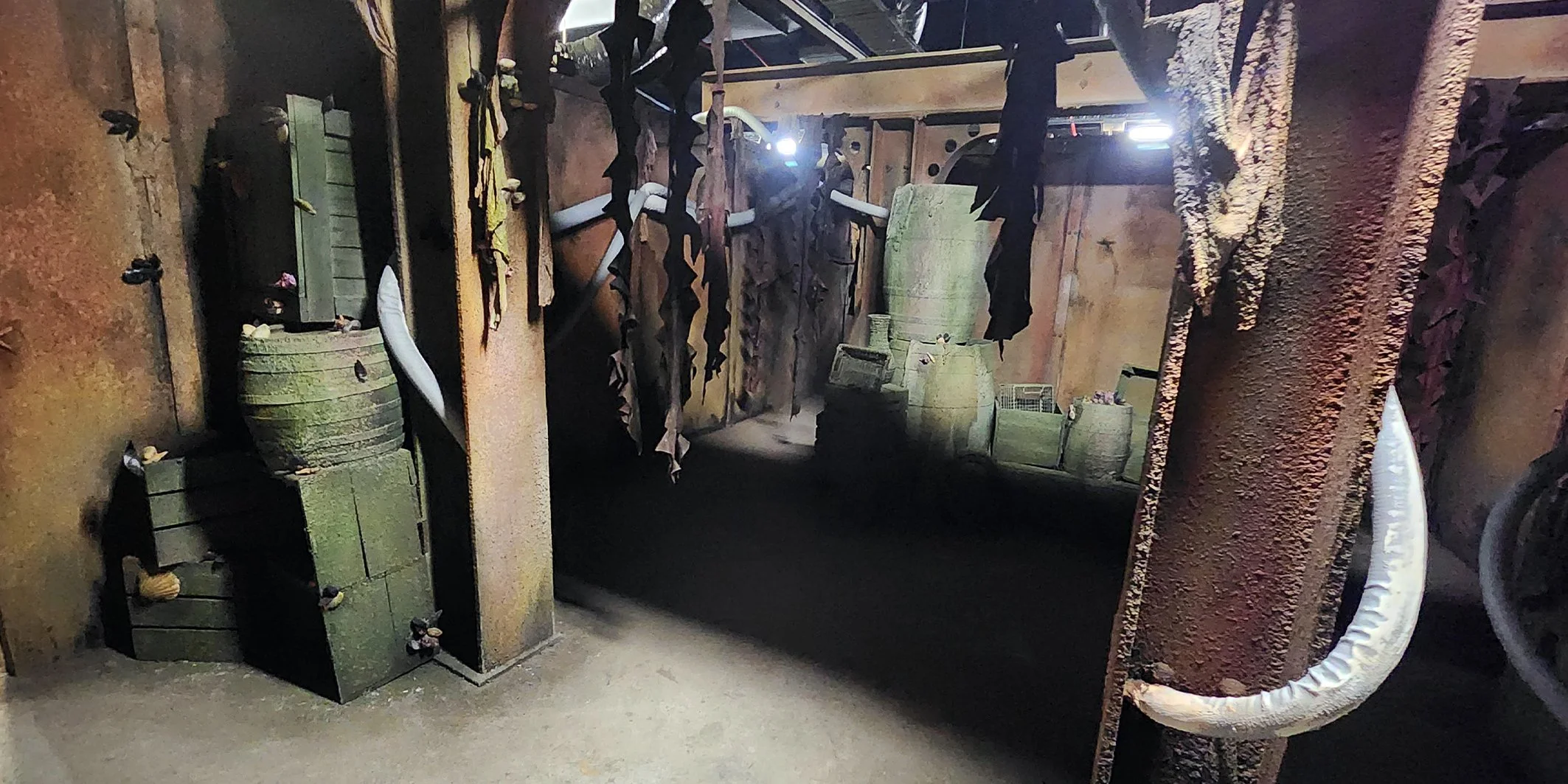 A dimly lit room with rustic decor, containing green wood crates, large ceramic pots, and a branch decorated with black leaves hanging from the ceiling. The walls are textured with a reddish-brown color, and there are various objects and furniture scattered throughout the space.