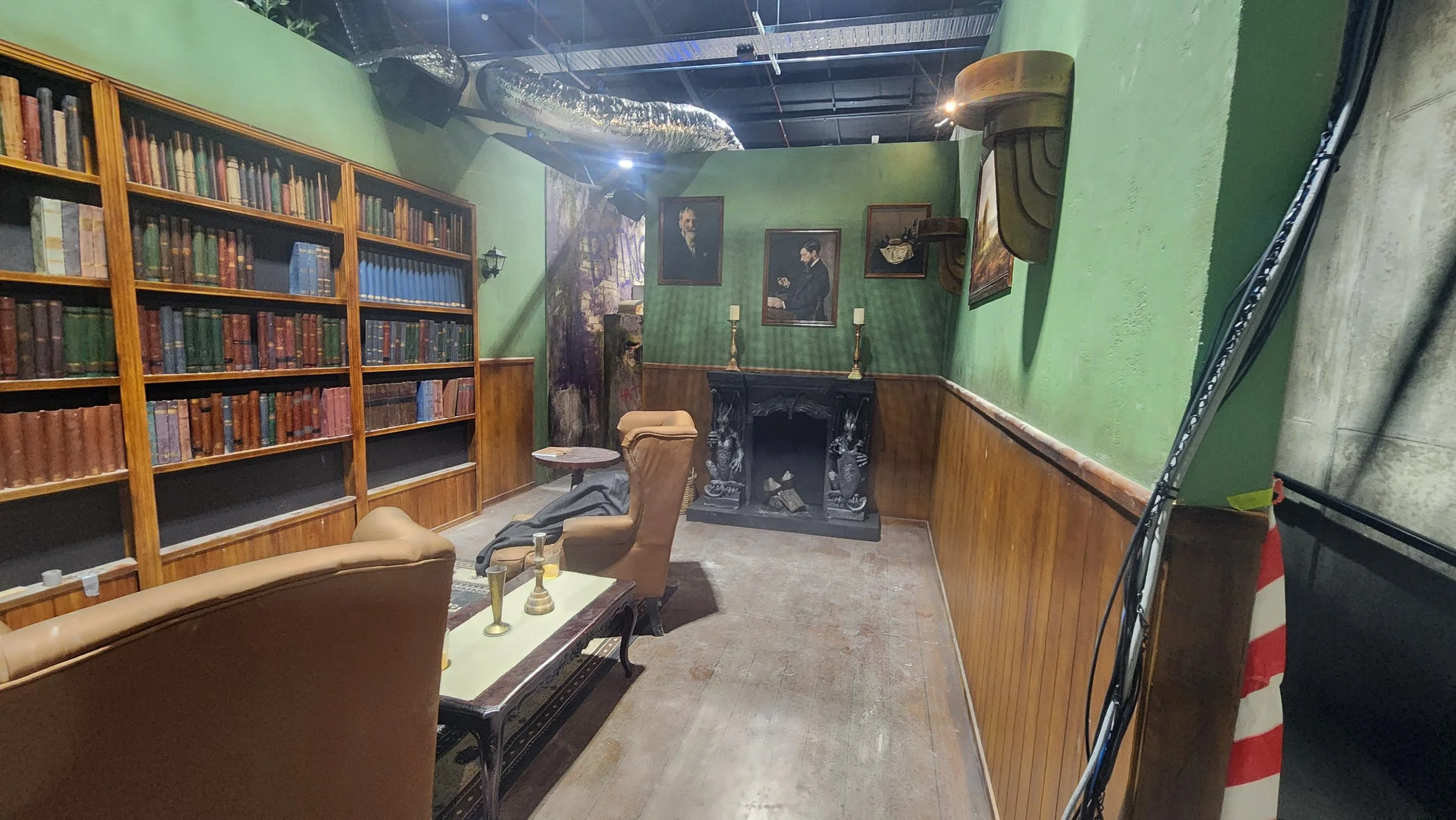 A cozy room with green walls, wooden bookshelves filled with books, a black ornate fireplace, and several chairs and a coffee table with decorative items. Portrait paintings hang on the wall, and there is a large silver duct on the ceiling.