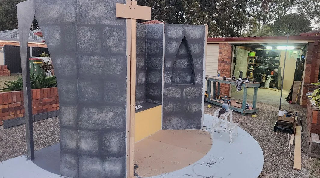 Construction of a small outdoor structure that resembles a castle, with gray stone-texture walls and arched window openings, in a backyard with a brick fence and a garage in the background.