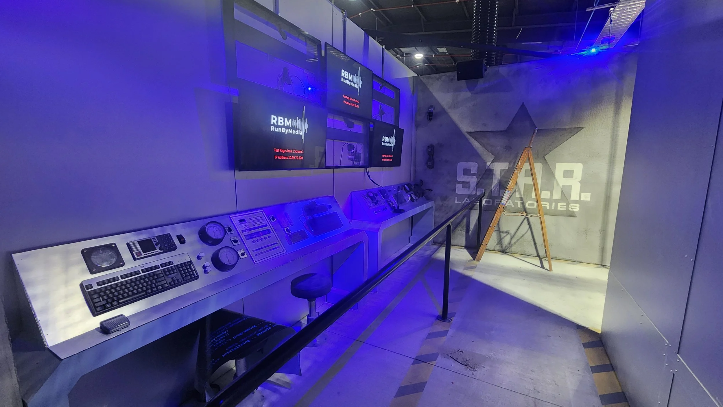 An electronic control panel with a keyboard, dials, and buttons on a stainless steel counter in front of multiple monitors, a ladder, and a wall with the Star Laboratories logo.