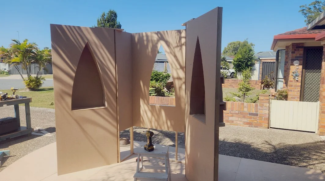 A woodworking project in progress with large beige colored panels featuring arched and rectangular cutouts, set up outdoors in a residential backyard with tools and a drill visible.