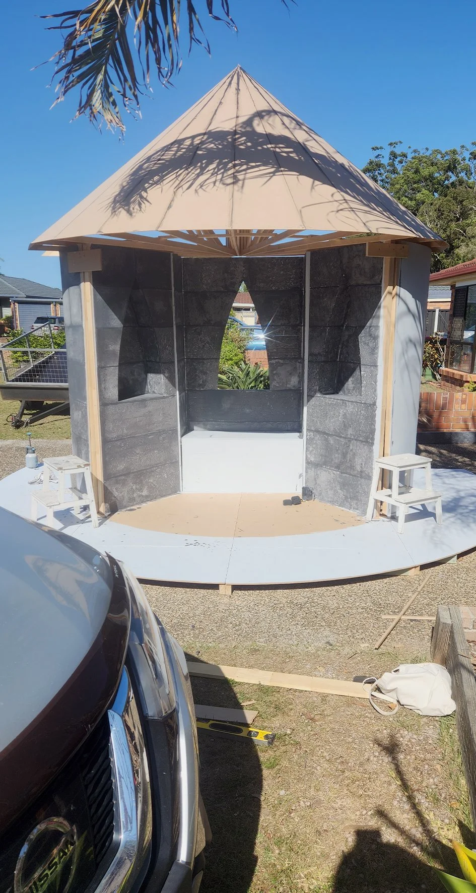 A small outdoor stage or shelter under construction, with a circular wooden platform and a tent-like beige canopy. There are two small white stools on either side and tools and materials nearby. The background shows trees and a residential neighborhood.