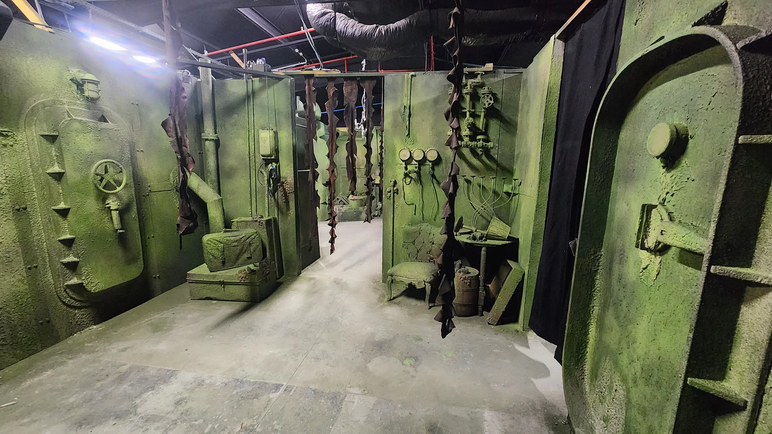 A room painted entirely in green with various old industrial or mechanical equipment, gauges, pipes, and hanging fabric strips, giving an industrial or steampunk aesthetic.