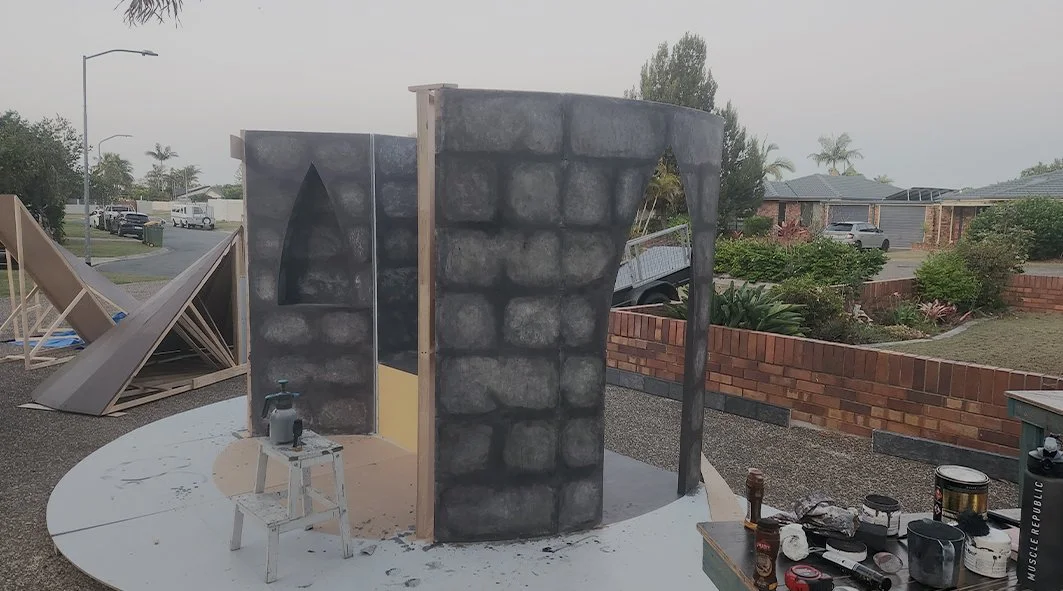 A partially constructed outdoor rock climbing wall with painted faux stone design, positioned on a circular base in a residential backyard
