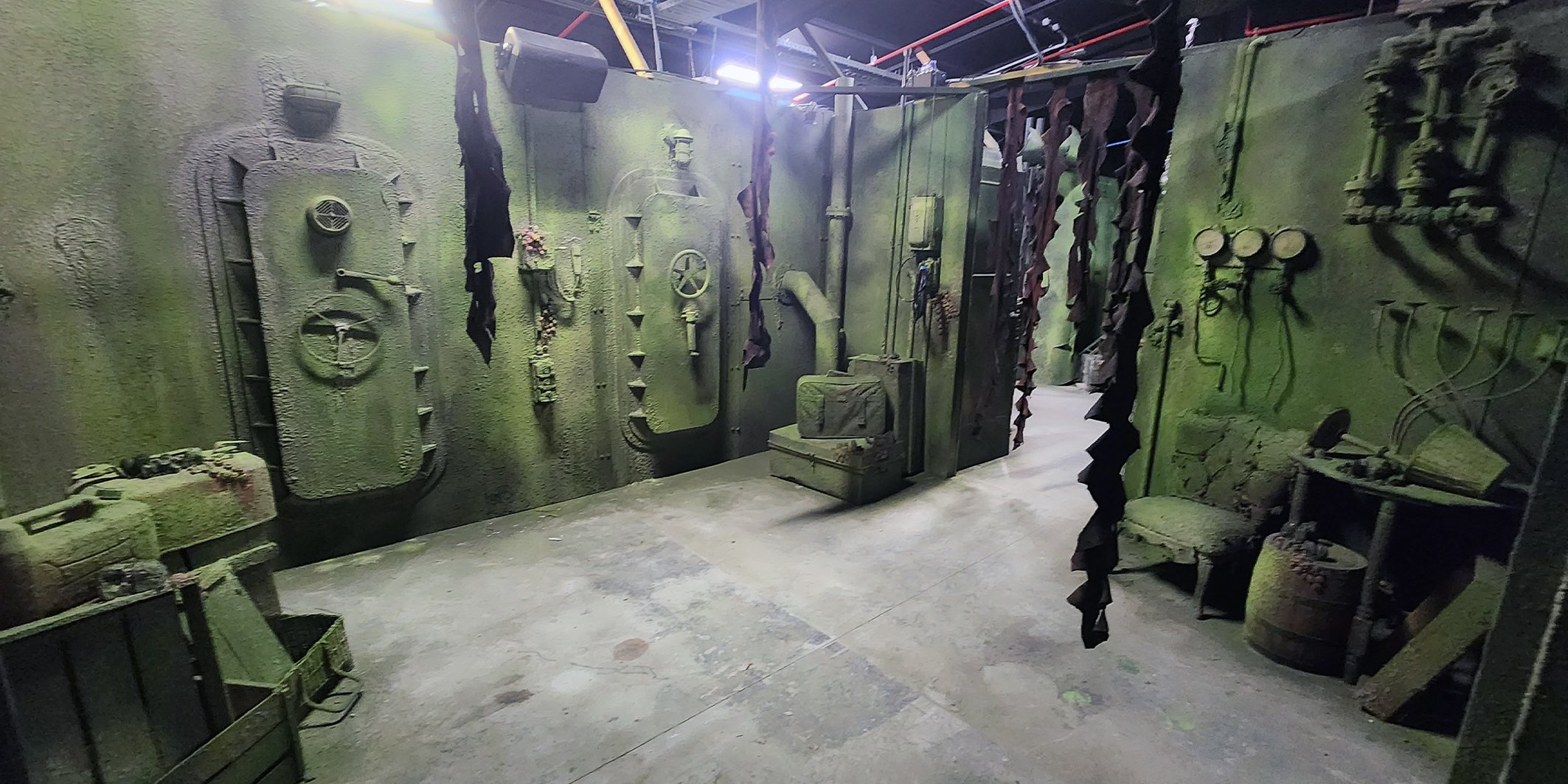 A room designed to look like a military bunker, with olive green textured walls, vintage military equipment, and clothing items hanging on the wall, including a chair, a trunk, and various gauges and valves.
