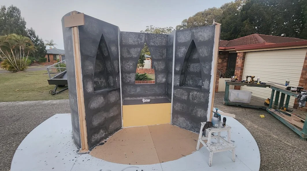 Partial construction of a Gothic-style wall with pointed arch openings, painted black with simulated stone texture, set on a circular base outdoors.