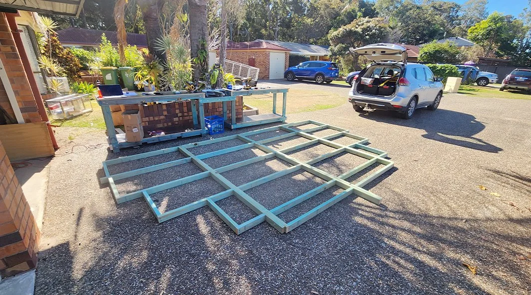 Unassembled outdoor wooden frame laid on a driveway near a screened porch with gardening plants, gardening tools, and a workbench, with parked cars and houses in the background.