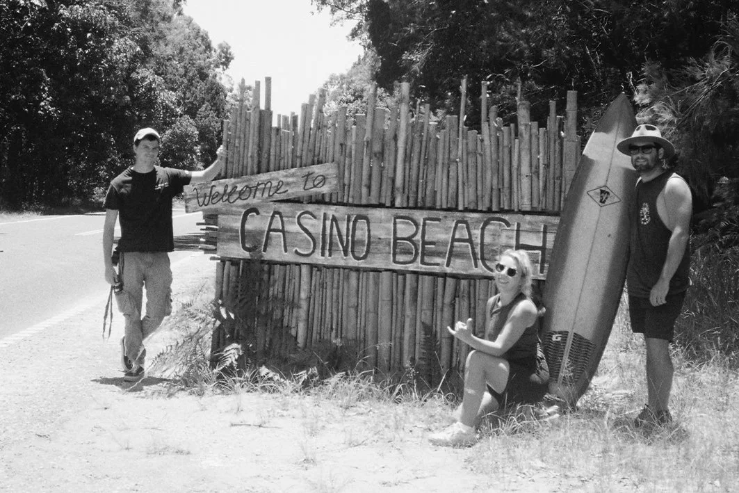 Three people, two men and a woman, at Casino Beach with a wooden sign that says "Welcome to Casino Beach". One man holds a surfboard, and the woman is sitting on the ground wearing sunglasses.