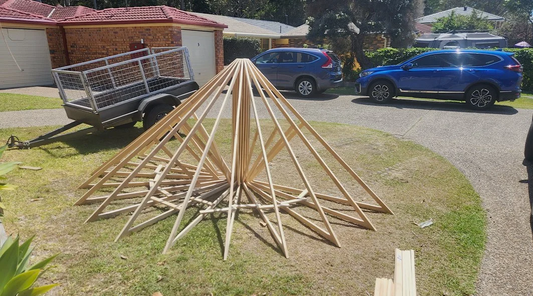 Wooden frames in a cone shape set up on a lawn, with trailers and parked cars in the background.