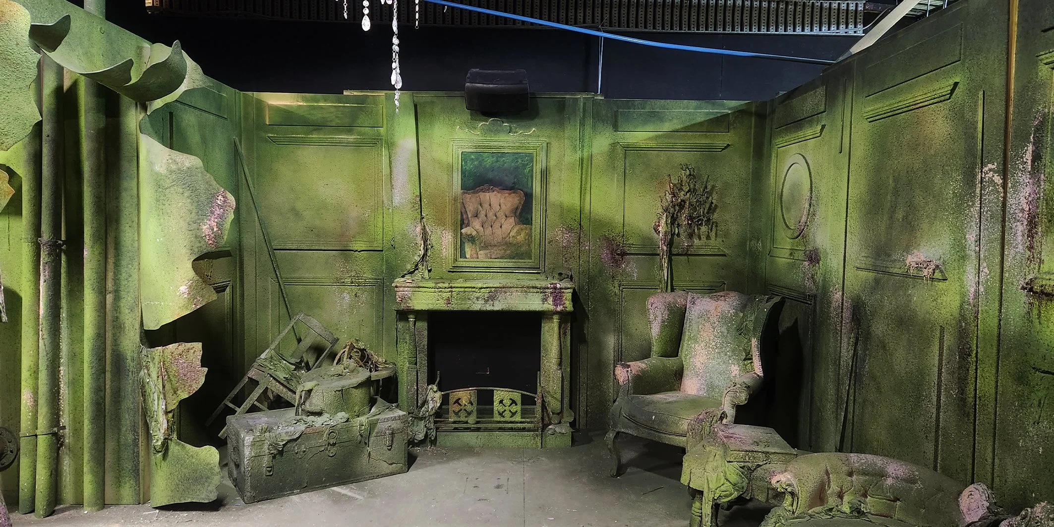 An old, abandoned room with green walls and furniture, covered in moss and mold. There are damaged chairs, a fireplace with a broken mirror above it, and debris scattered on the floor.