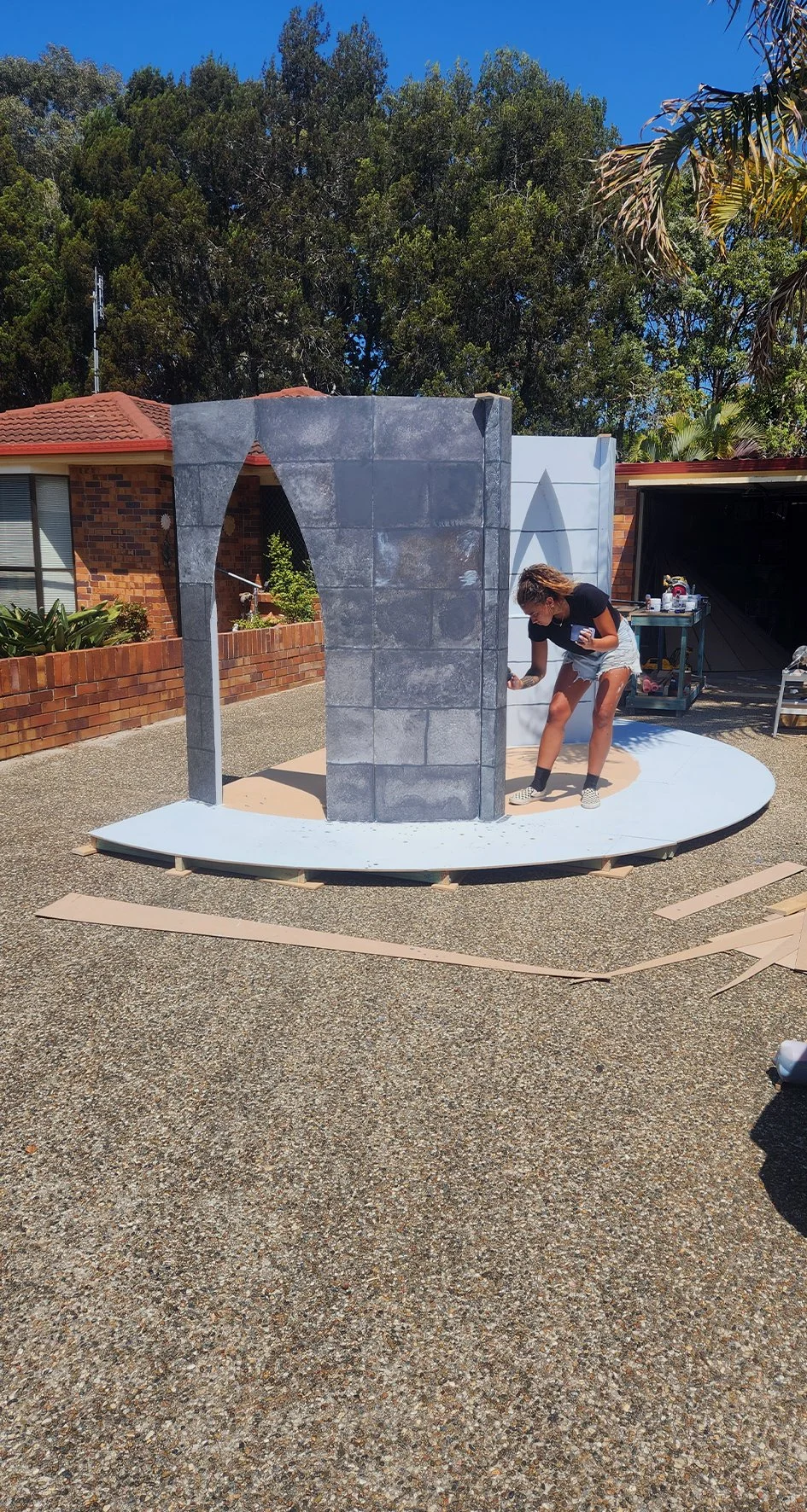 A woman painting a small set design resembling a stone structure with an arch and a wall, outdoors on a sunny day.