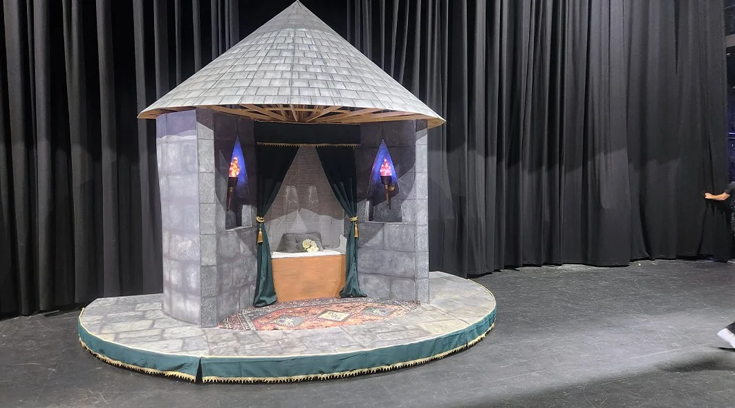 Miniature theater set resembling a castle with a small bed, curtains, and decorative elements, on a stage with black curtains in the background.