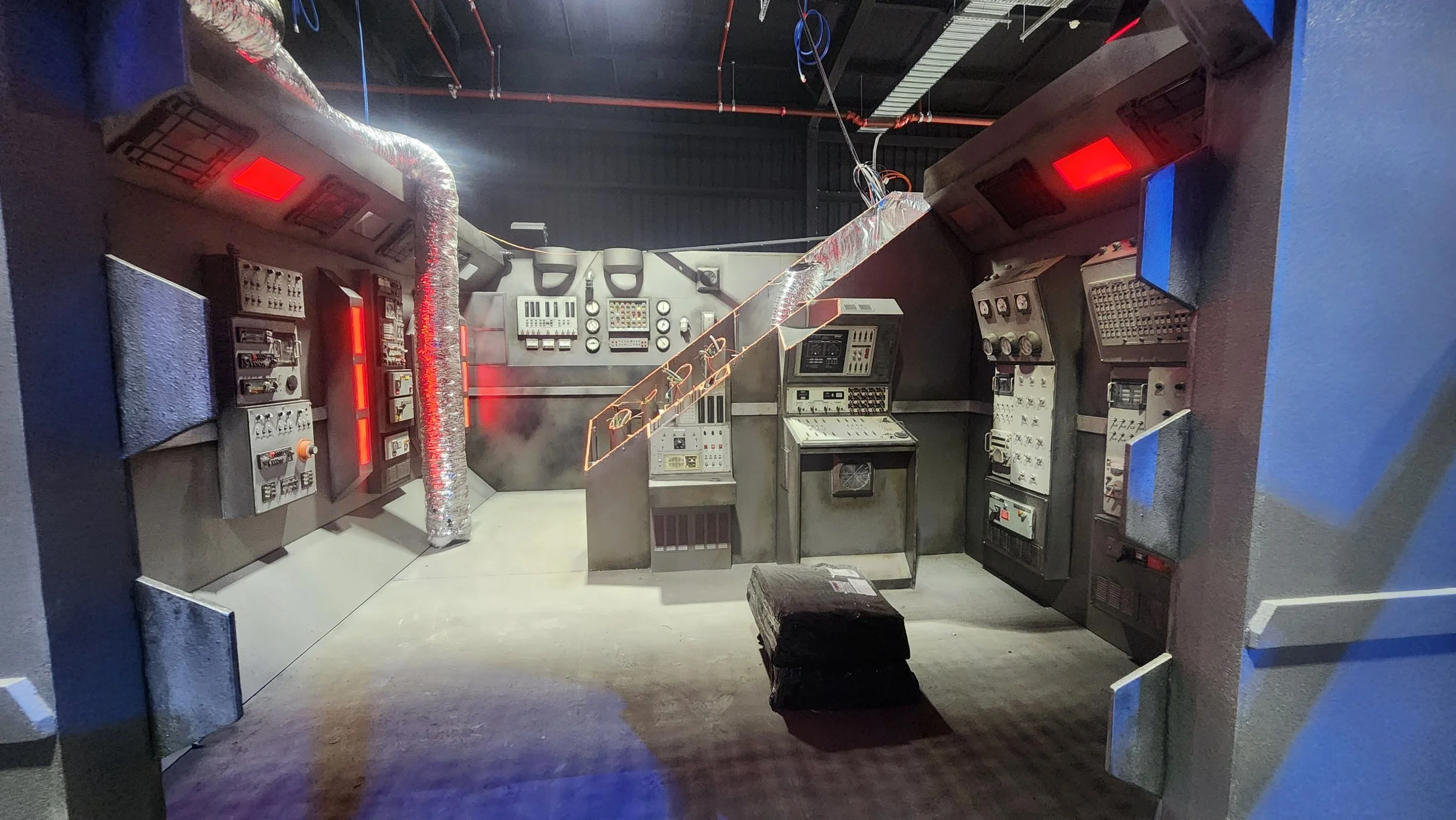 An industrial-style room filled with old electronic equipment and control panels, lit with red and blue lights, with exposed ductwork and industrial piping on the ceiling.