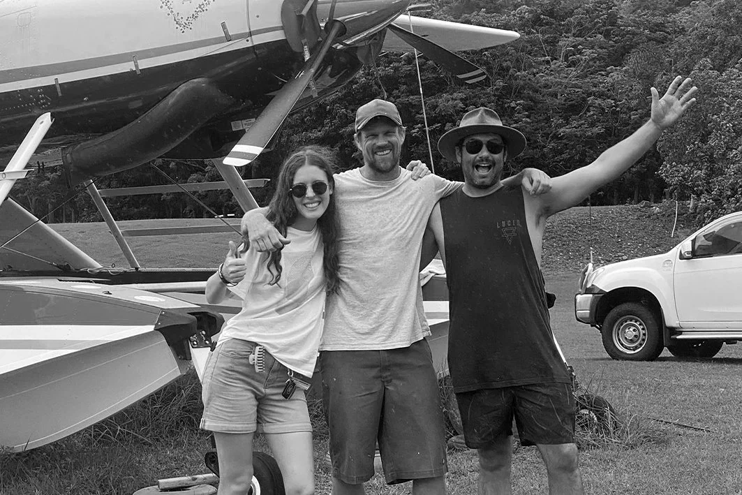 Three friends stand in front of a floatplane at an outdoor location, smiling and posing for the camera, with trees and another vehicle in the background.