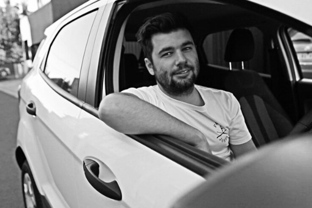 A man with a beard and dark hair smiling and sitting in a car with his arm resting on the window frame, outside on a street.