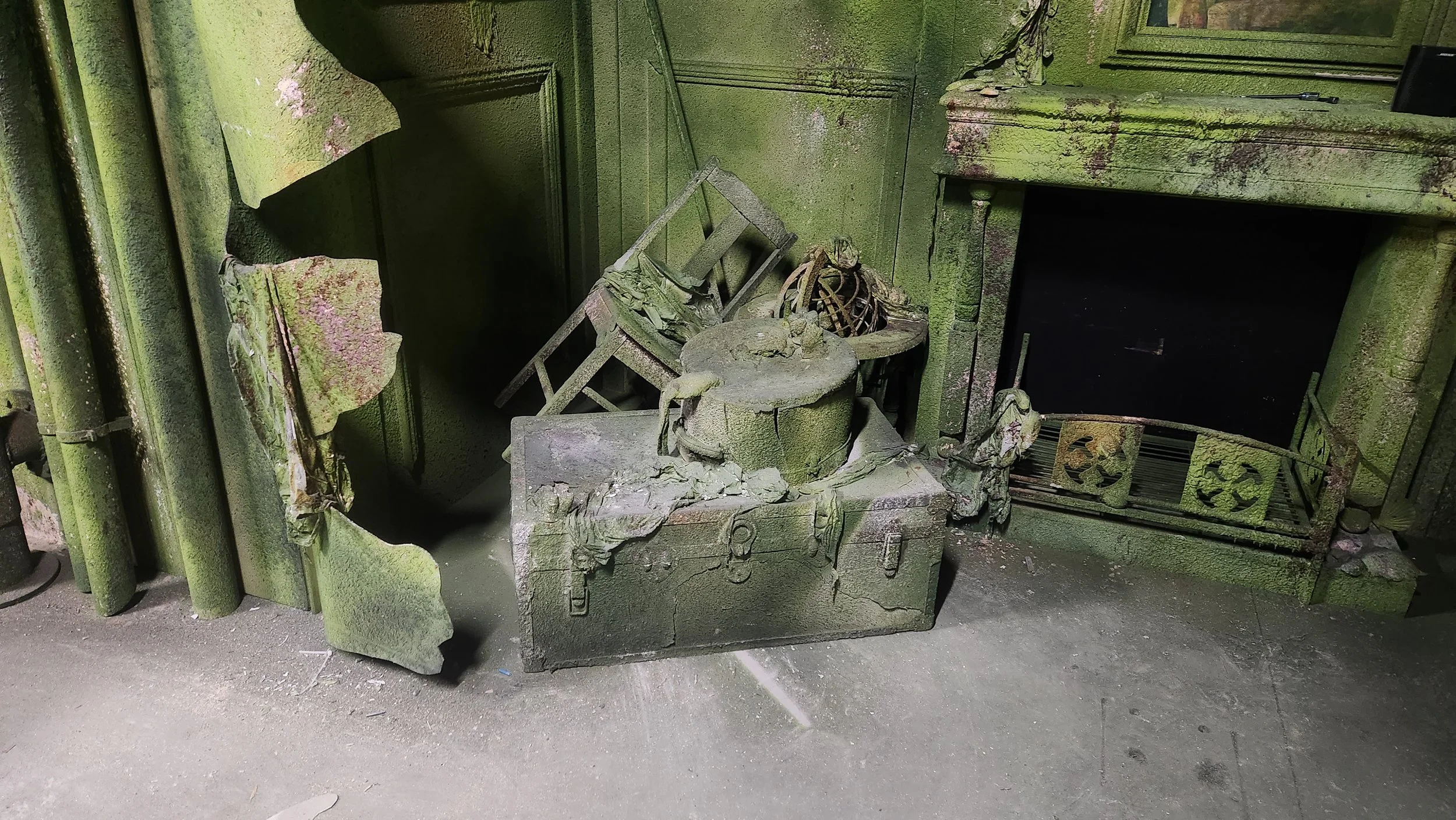 A room covered in thick green moss and mold, featuring a damaged stone fireplace with an open firebox and a pile of debris including a small sculpture of a dog, various broken furniture pieces, and an old, tilted chair.