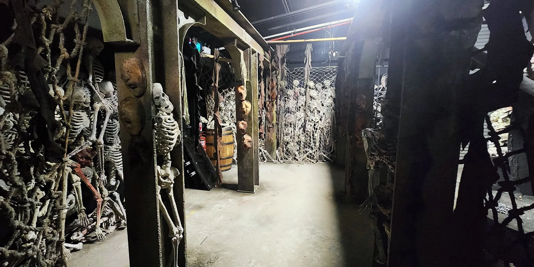 A storage room filled with skeletons and costumes hanging on racks. There are barrels and miscellaneous items, with some skeletons in cages and others arranged along the walls. The room has a dark industrial environment.