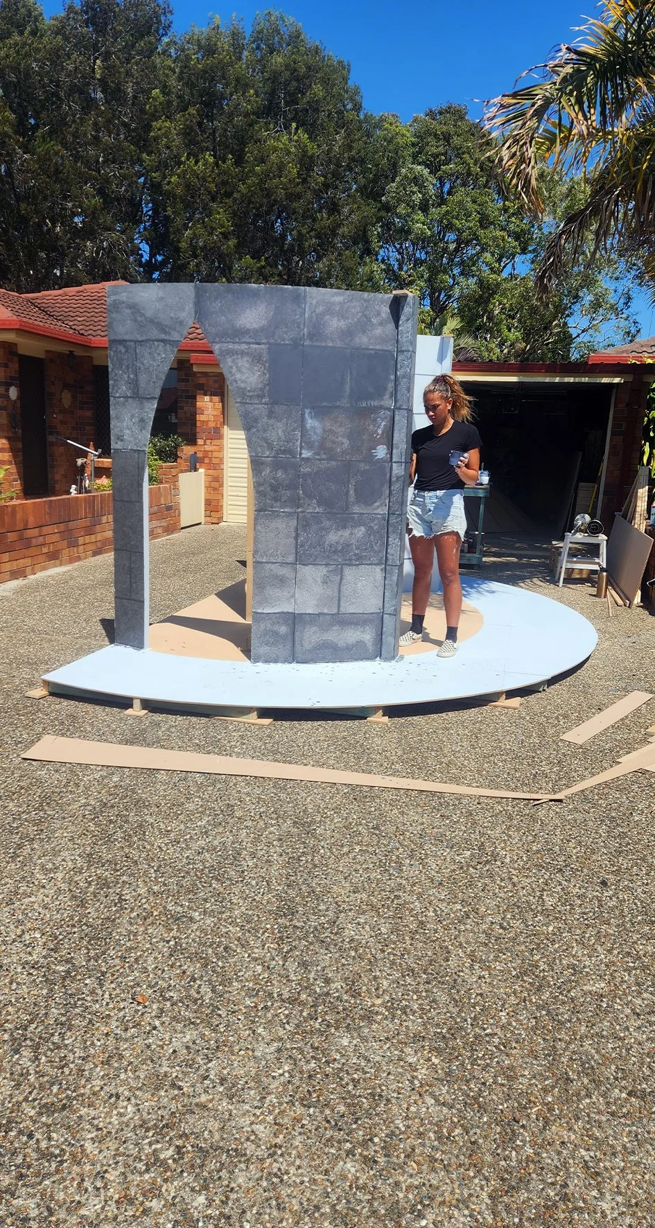 A woman standing near a partially constructed circular structure with a brick wall exterior, on a textured driveway under a blue sky with trees in the background.