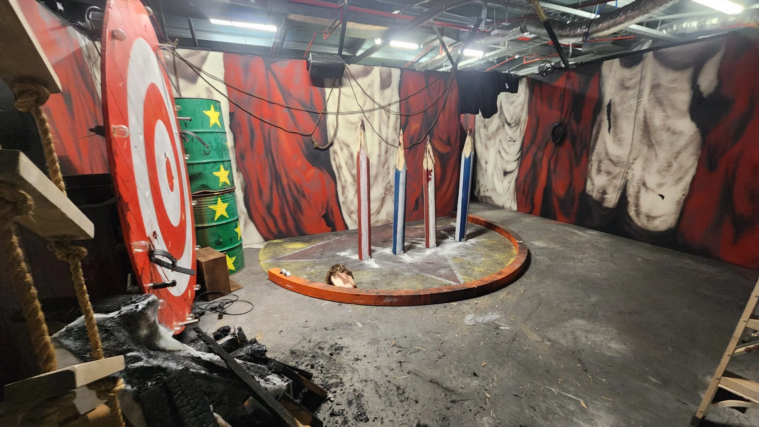 An indoor space with painted walls featuring a tiger and other abstract designs. There are large drums, ropes, and wooden structures, suggesting an obstacle course or themed activity space. The floor is covered with dirt and debris, and there is a circular area with a person’s head barely visible, possibly part of a game or activity setup.