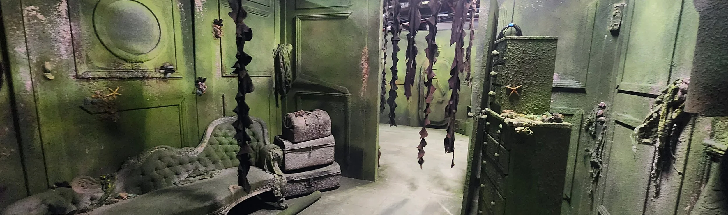 An abandoned, grimy room with green paint peeling off the walls, old damaged furniture including a tufted sofa, a small chest, and a dresser, cluttered with debris, dried sea stars, and dried seaweed. Tattered, dark curtains hang from the ceiling, and the overall scene appears neglected and decayed, giving an eerie, haunted atmosphere.