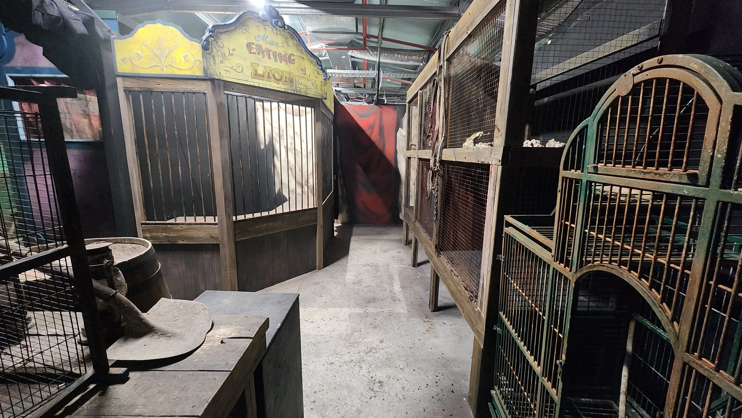 A storage room with animal cages and enclosures, including wooden and metal cages, some containing small animals, a feeding trough, and a sign that reads 'Eating Lion' in the background.