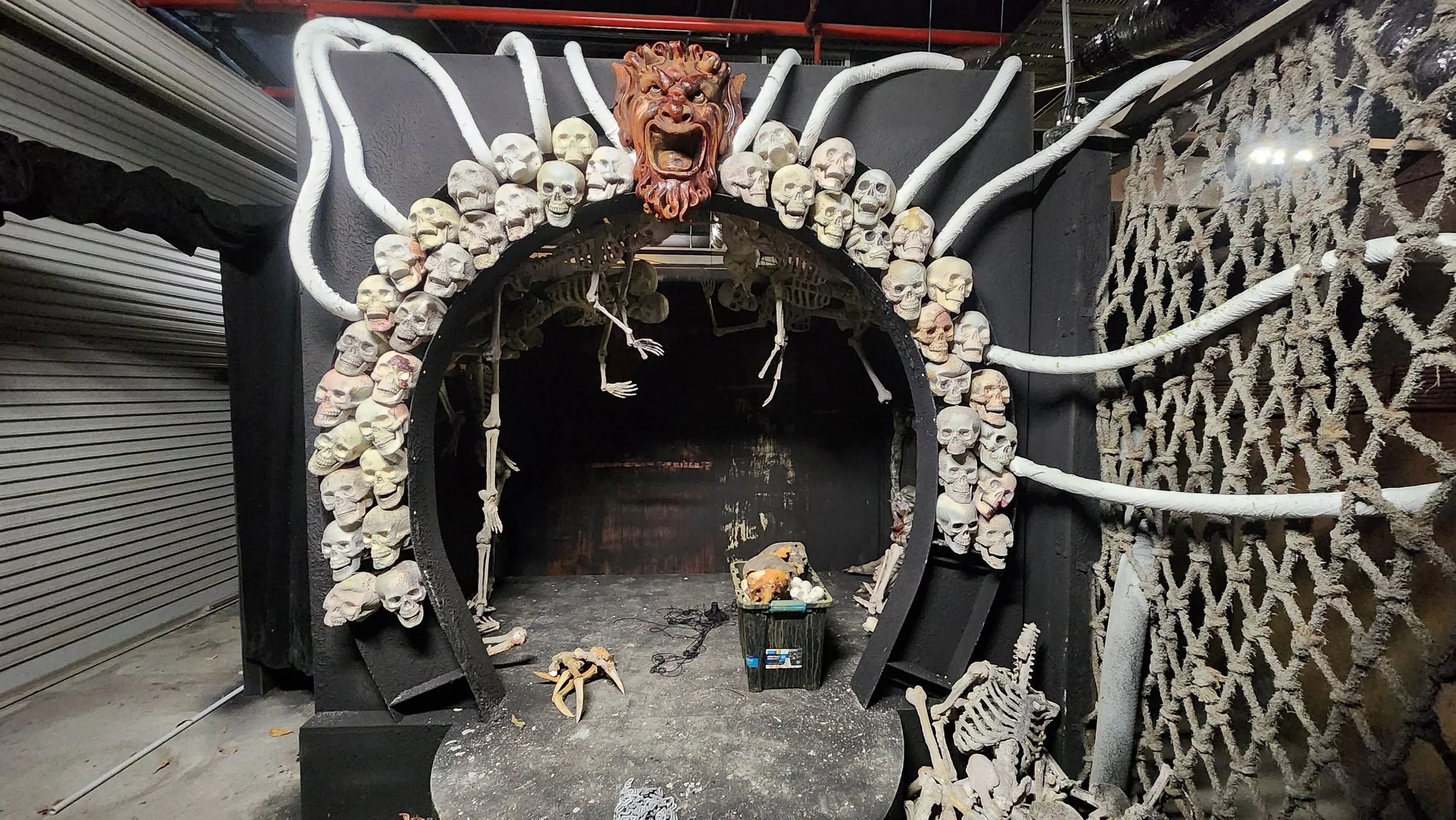 A Halloween-themed display featuring a circular arrangement of skulls and skeleton bones. A fierce lion's head with an open mouth is mounted at the top center, surrounded by white plastic or fabric tubes resembling spider legs or tentacles. The background is black with some construction materials and tools on the ground, and parts of a gray roller shutter on the left and a decorative mesh with skulls on the right.