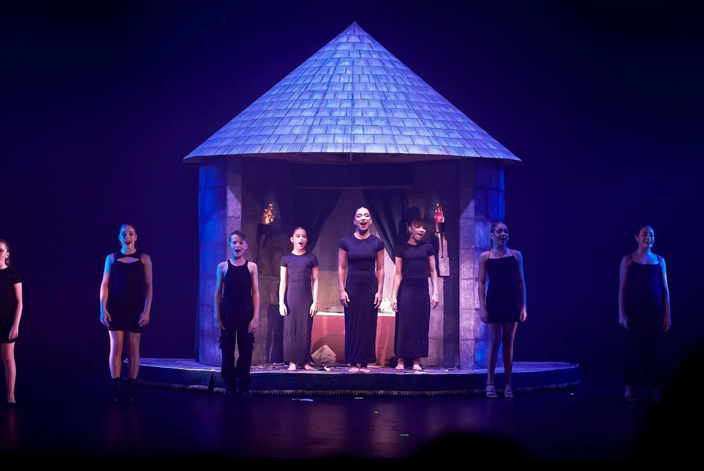 Theater performance with seven young girls in black dresses singing on stage in front of a faux castle backdrop with blue and purple lighting.