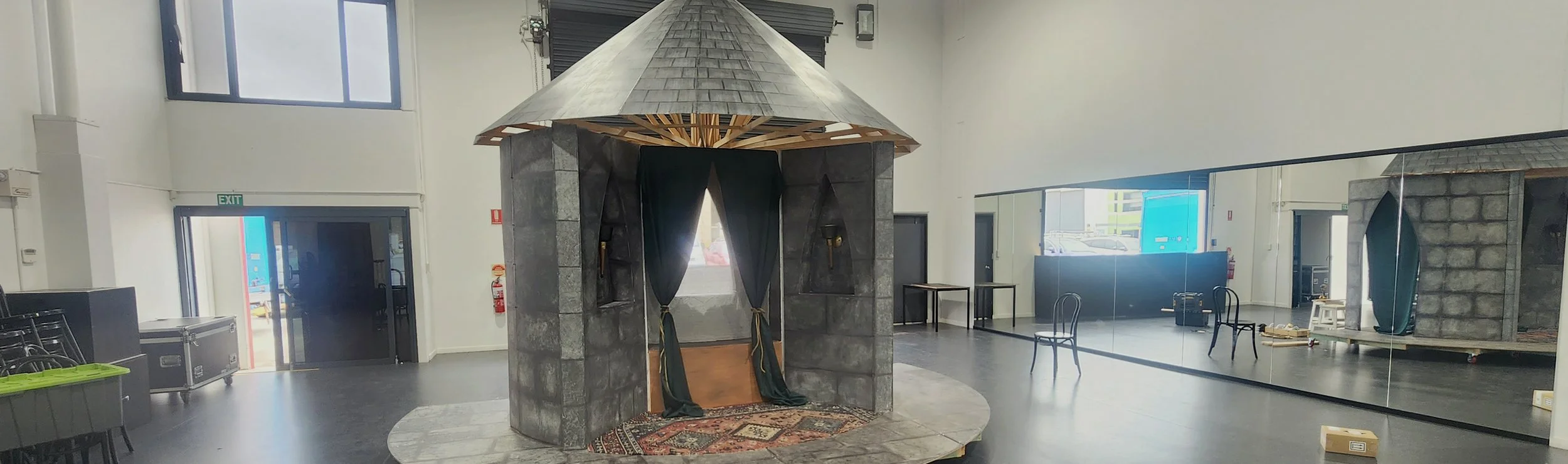 An indoor theater set with a small castle-like structure made of stone-like material, featuring a peaked roof and enclosed stage area with curtains. The space has a black floor, mirrored wall on the right, and various props and chairs around.